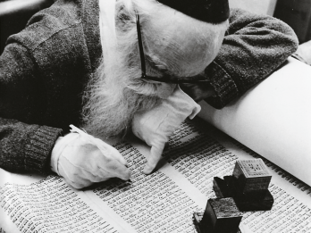 Photograph depicting man in kippah, gloves, and glasses and holding a pen examining a Torah scroll upon which sits two tefillin boxes.  