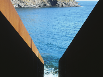 Photograph of metal sculpture in the shape of a V through which the ocean and rocky outcropping beyond is visible. 