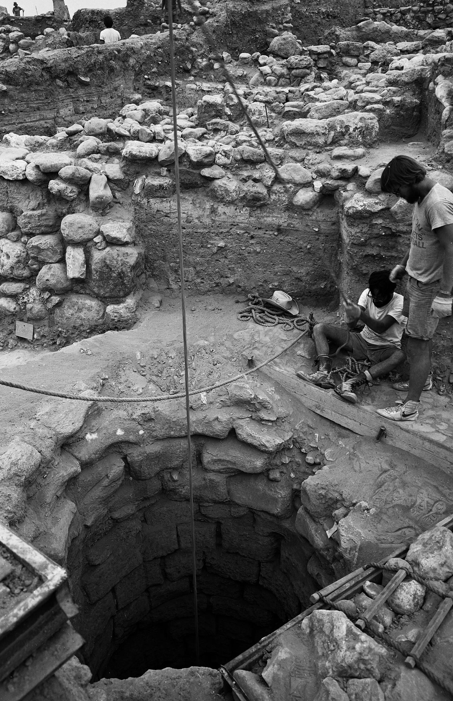 Photograph of stone well with rope system and two figures standing on the side. 