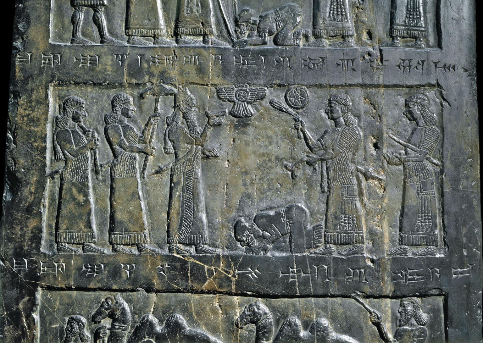 Relief of five standing figures carrying objects, and another figure prostrate in the middle of figures, surrounded by rectangular border with Akkadian inscription. 
