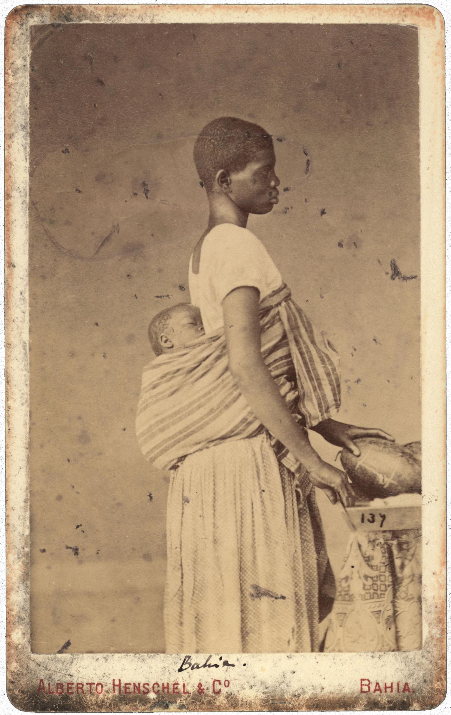 Full body profile photograph of person in profile with baby in shawl on her back.