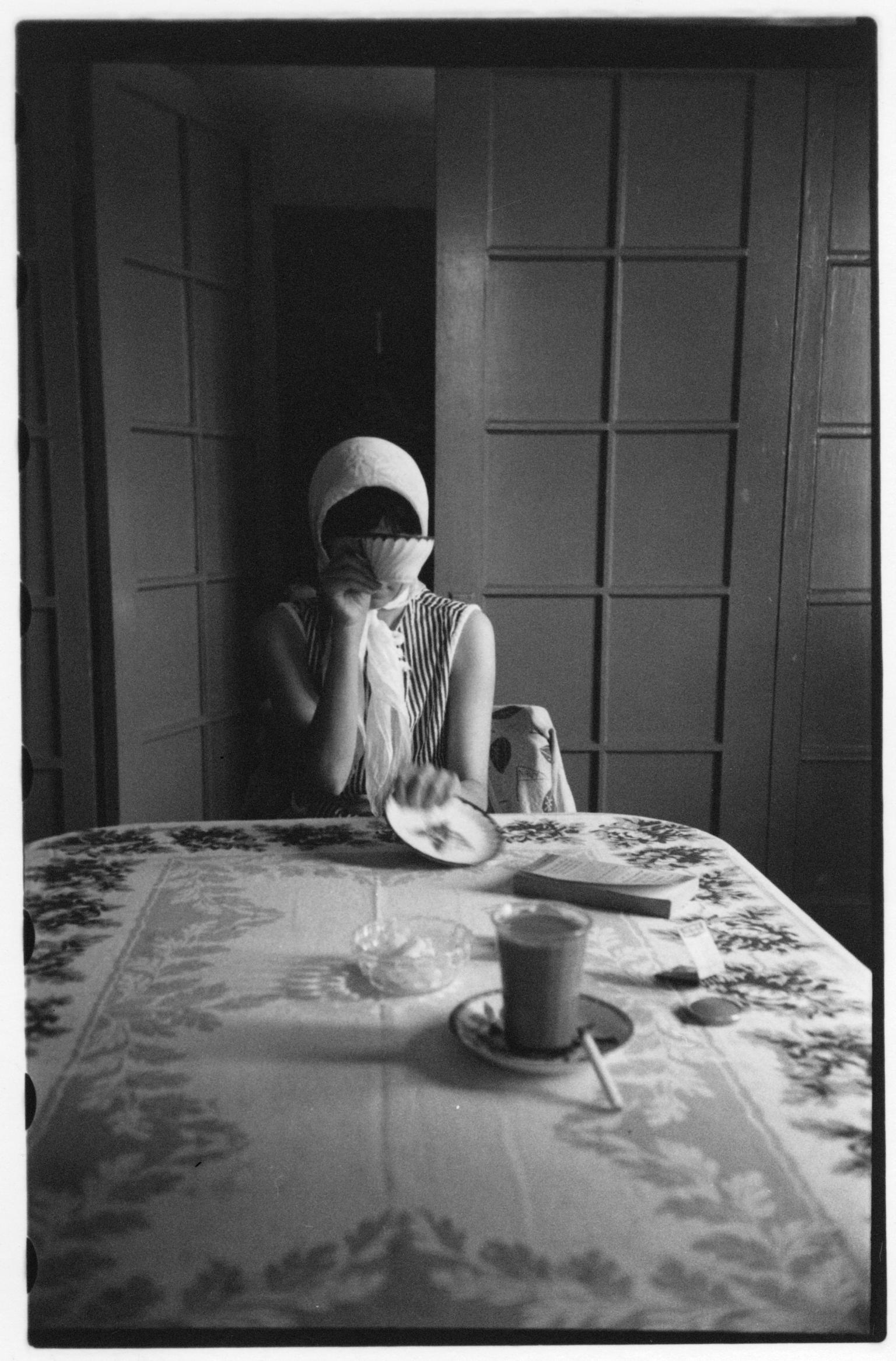 Photograph of woman sitting at the end of a table in front of open doors, wearing a scarf and covering her face with tea cup.