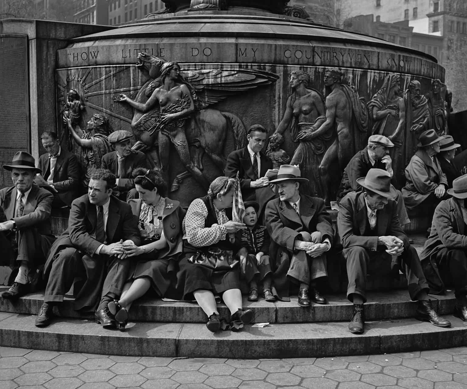 Photograph of men, women, and children seated on the steps of a large public monument with nude classical figures and a winged horse atop a circular base.
