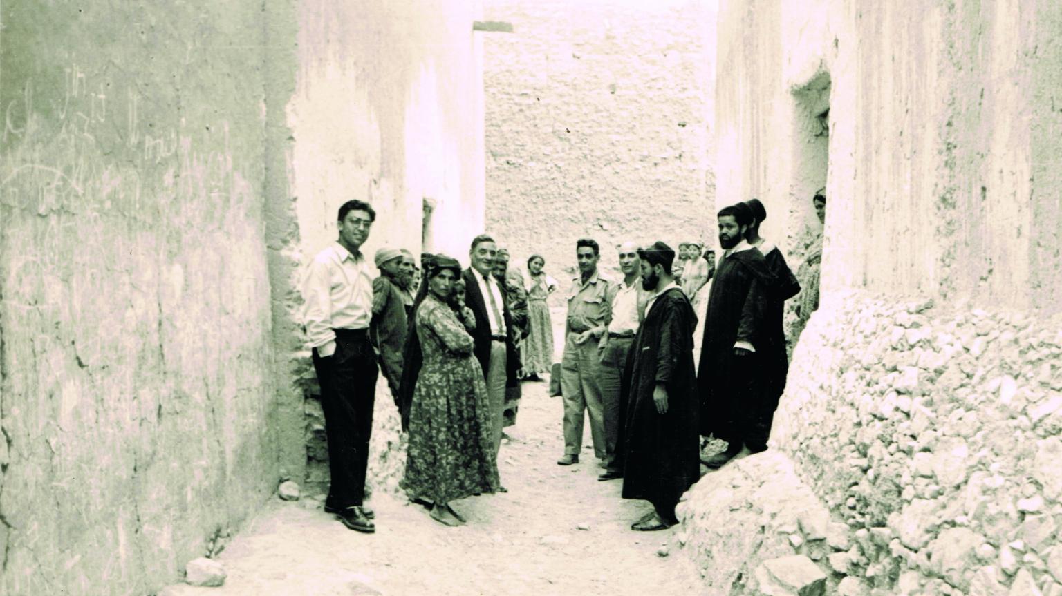 Photograph of men and women in an alley, most facing camera while others look away.