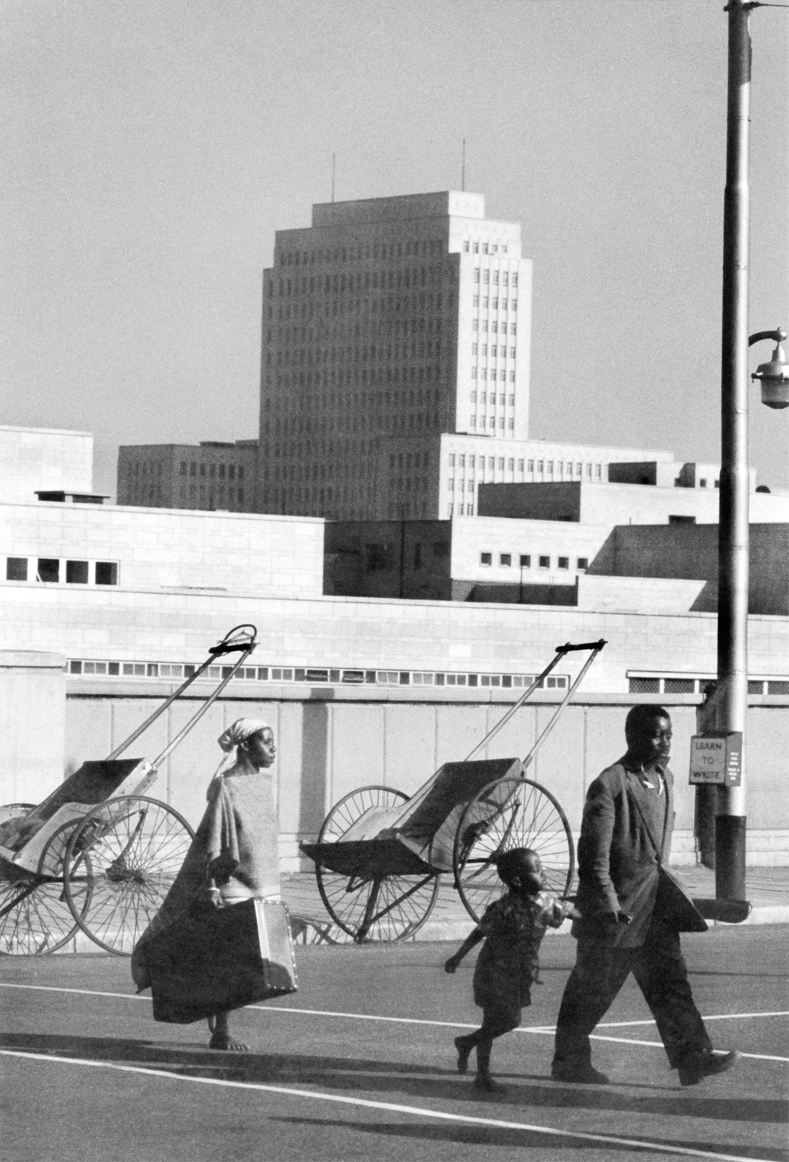 Photograph of man, woman, and child walking down city street with high-rise building and two rickshaws in background.