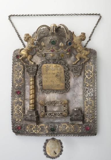 Shield with decorative border inlaid with semi-precious stones and adorned with lions and crown at top, column on left and plaque in middle with Hebrew text. 