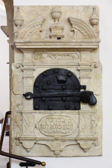 Carved wall unit with decorative borders and crowns, locked metal box in middle, and Hebrew text underneath. 