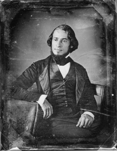 Portrait of man in suit facing slightly angled, seated with his arm resting on the table.
