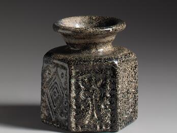 Photo of a dark glass vase with a wide, flared rim and hexagonal base, featuring geometric patterns.