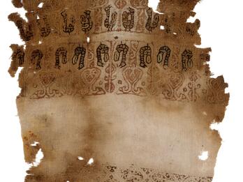 Damaged fragment of fabric with decorative patterns and symbols in dark ink and faded color, mainly red, toward the top and in one additional stripe at the bottom.