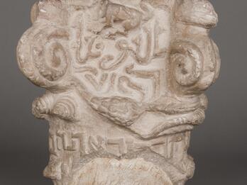 Worn carved stone capital featuring spirals, patterns, flora and fauna, and lines of Arabic and Hebrew lettering.