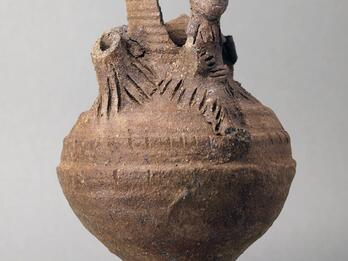Earthenware vessel with a round body and flared base, with sculpted elements, some broken off, at the top, including a humanoid figure.