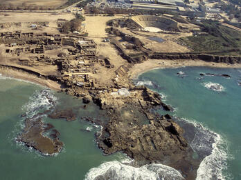 Aerial view of ancient ruins along a coastline, with stone structures and an amphitheater near the sea, surrounded by water and rocky shores.