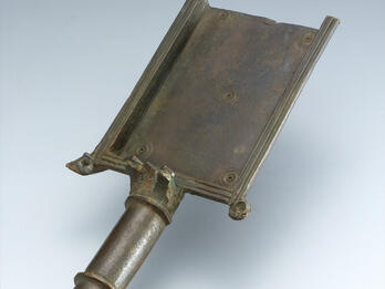 Ancient bronze shovel with a rectangular surface and a tapered cylindrical handle.