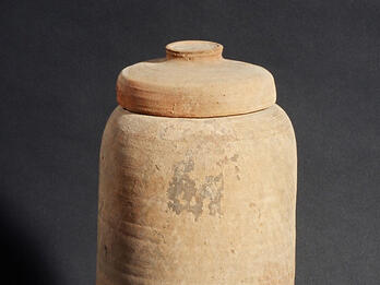 A tall, cylindrical clay jar with a rounded lid, standing upright against a dark background. The surface appears rough and slightly uneven, with some discoloration and markings.