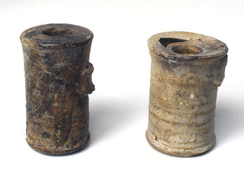 Two ancient cylindrical jars with small openings at the top, both weathered and partially damaged, standing upright.