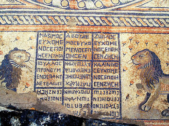 Ancient mosaic featuring two lions facing a central grid with Greek letters, surrounded by decorative patterns. The scene appears weathered, with part of the mosaic damaged in the lower left corner.