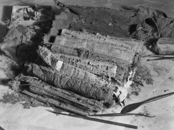 A close-up black-and-white photo of fragile, frayed rolls of papyrus. Restoration tools and a tray with small fragments lie nearby on a work surface.