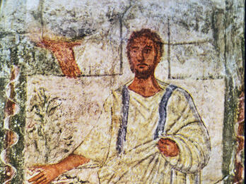 Ancient fresco shows a bearded man in a white robe with blue stripes, standing beside a tree, extending one arm outward. The background features simple landscape shapes, and a wavy border surrounds the scene.