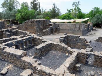 Stone ruins with partial walls, including some with window-like openings, surrounded by trees and greenery; shade cloths are stretched above part of the site.