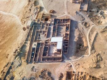 Aerial view of ancient ruins in a desert landscape, showing rectangular stone foundations, partially standing walls, and pathways winding around the site. The terrain is arid and rocky with sparse vegetation.