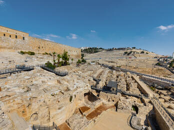 Ancient stone ruins and archaeological site near a large stone wall, with hills and scattered buildings in the background under a clear blue sky.