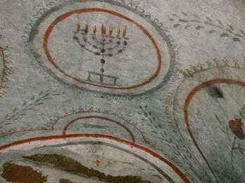 Ancient painting featuring a menorah with seven lit candles inside a bordered circle, surrounded by decorative patterns and arches on a stone surface.