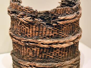 A worn, cylindrical woven basket with a broken, jagged rim and thick, braided bands around its body, displayed on a plain white surface.