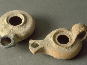 Two ancient, round clay oil lamps with spouts and central openings rest on a plain surface. The lamps are worn and show earthy tones.