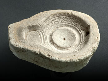 Ancient stone mold for an oil lamp with a circular reservoir, intricate geometric carvings around the rim, and a spout for the wick.