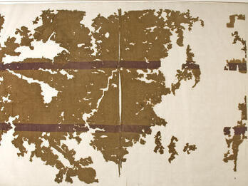 A brownish, ancient textile fragment with dark red horizontal bands on a cream background, showing heavy wear and missing sections.