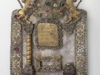 Shield with decorative border inlaid with semi-precious stones and adorned with lions and crown at top, column on left and plaque in middle with Hebrew text. 