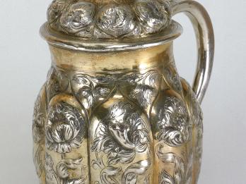 Silver pitcher with floral shapes and inscribed designs, and shield on lid with Hebrew inscription. 