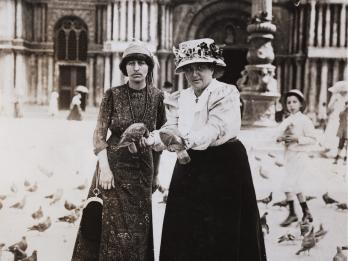 Two women in hats surrounded by pigeons in city plaza, with ornate doorway behind them.