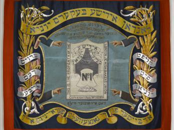 Banner with four arms pointed toward a crest in the center featuring a loaf of bread and English text, framed by Yiddish text and sheaves of wheat.