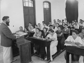 Photograph of classroom with teacher at podium and men and women at rows of desks.