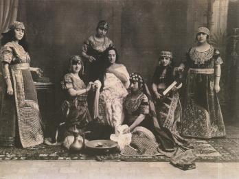 Photograph of several women in dresses and headdresses with rug and cooking implements on the floor.