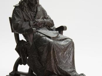 Sculpture of seated figure with blanket draped over legs and book in lap.