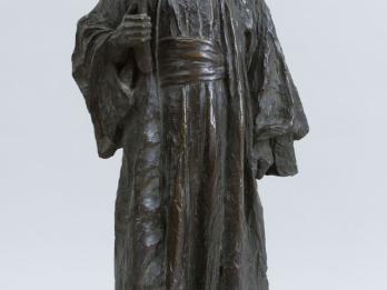 Sculpture of a figure in robe and hat, carrying a book.