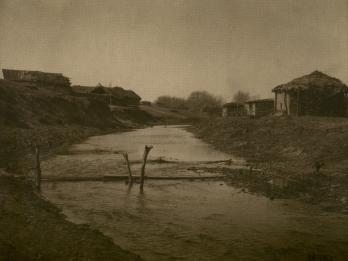 Photograph of landscape with creek lined with small huts.