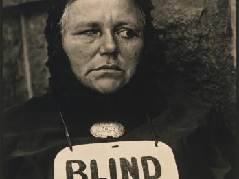 Photograph of woman wearing a sign around her neck that reads "BLIND."
