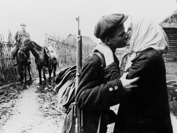 Photograph of mother kissing her son, who has a gun and a backpack, in foreground, while another soldier waits in the background on his horse holding the reins of another horse. 
