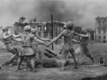 Photograph of children holding hands in a circle and dancing around an alligator statue as a city burns in the background.