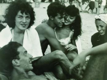 Photograph of five people sitting closely together, with one lying down, in a circle on the beach. 