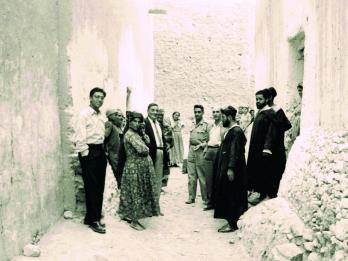 Photograph of men and women in an alley, most facing camera while others look away.