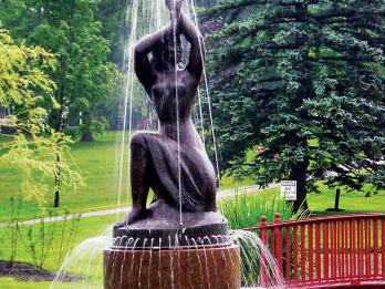 Bronze sculpture and fountain in the shape of a partially nude woman, resting on one knee and raising her arms above her head, set in a park with trees and a bridge in the background. 