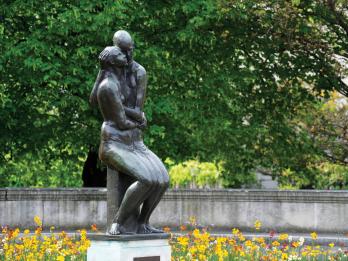 Bronze sculpture of young couple, seated side-by-side and cheek-to-cheek in a garden. The ground is covered by leaves and there are many trees in the background beyond a low wall.