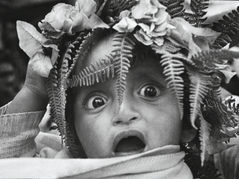 Photograph of a wide-eyed baby with mouth open and ferns and flowers on their head. 