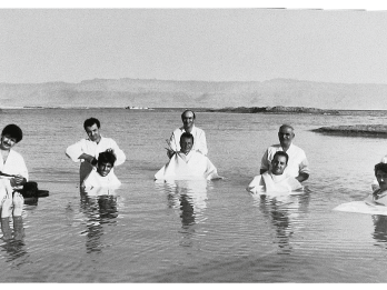 Photograph featuring seven barbers standing in the ocean and cutting hair of seven men floating in front of them. 