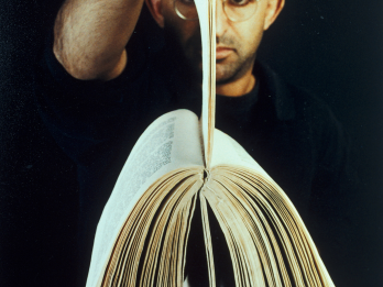 Photograph showing man facing camera and holding up a book by its pages.